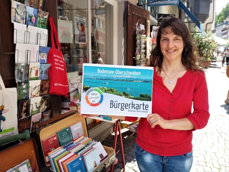 Vor einem innenstädtischen Ladengeschäft hält eine Frau mit rotem Oberteil eine vergrößerte Abbildung der Bürgerkarte Bodensee-Oberschwaben frontal in die Blickrichtung.