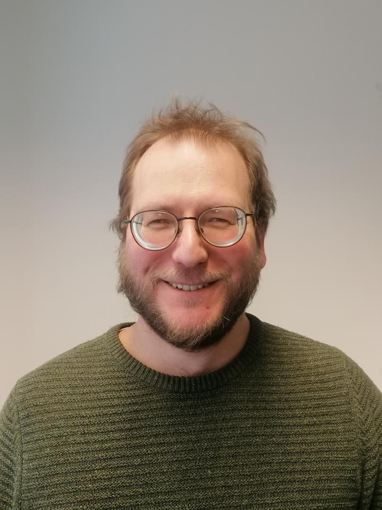 Ein Porträtlichtbild von Jonas Buchhardt. Er lächelt in Richtung der Betrachterperspektive, trägt Kurzhaarfrisur, Brille, Bart und einen olivgrünen Pullover.