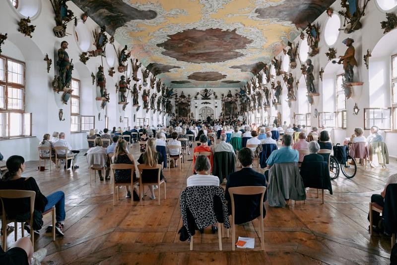 Ein historischer Schlosssaal mit Deckenfresken und Figurenplastiken an den mit Fenstern versehenen Seitenwänden ist gefüllt mit einem einer Bühne zugewandten Publikum. Die Personen sitzen mit Abständen getrennt voneinander. Je zwei Stühle sind nebeneinander angrenzend gestellt. Am vorderen Ende des langen Saales ist ein Orchester platziert.