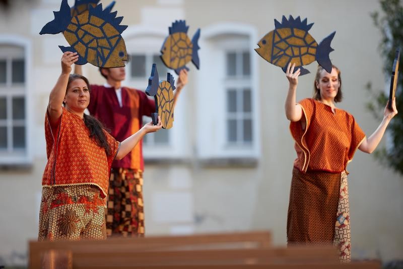 Das Bild zeigt draußen vor einem Gebäude drei Personen, zwei weibliche vordergründig, eine männliche hintergründig. Die Personen tragen traditionelle außereuropäische Kleidung in warmen Farbtönen von rot über orange bis terracottabraun, teilweise in gemustertem Dekor. Sie halten jeweils zwei bemalte Holzfische mit ihren Händen in die Höhe.