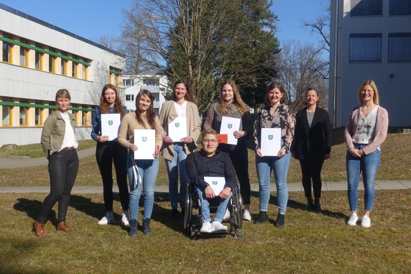 Das Bild zeigt die Absolventen und Absolventinnen der Ausbildung beim Landkreis. Auf einer Wiese vor einem Baum und Gebäuden stehen die Personen in zwei Reihen, der Fotoperspektive zugewandt.