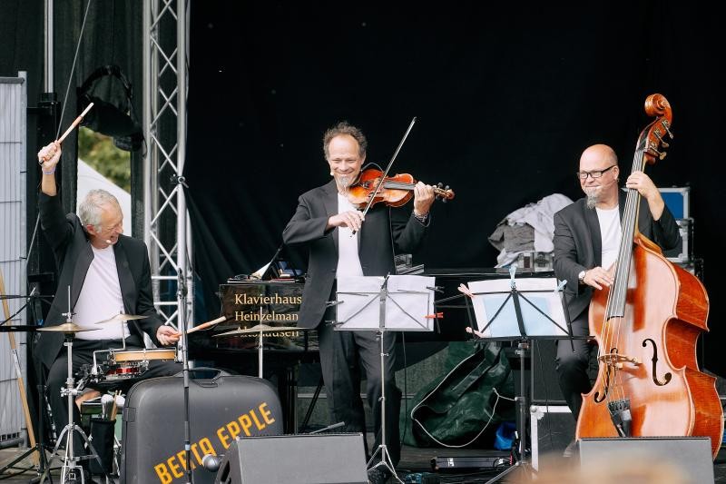Drei Männer mittleren Alters musizieren auf einer Bühne mit schwarzem Hintergrund. Sie tragen dunkle Jacketts und darunter weiße T-Shirts. einer spielt Cello, der zweie Geige, der dritte Schlagzeug.