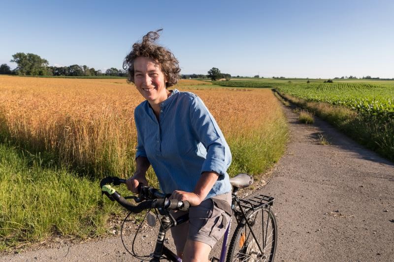 Eine Fahrrad fahrende Frau auf einem asphaltierten Feldweg entlang von Kornfeldern in sommerlicher Wetterlage.