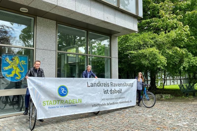 Drei Personen mit Fahrrädern halten gemeinsam ein langgezogenes Plakat  und stehen vor einem Verwaltungsgebäude des Landkreises Ravensburg. Auf dem Plakat ist das Logo der Aktion Stadtradeln und daneben der Text: Landkreis Ravensburg ist dabei.