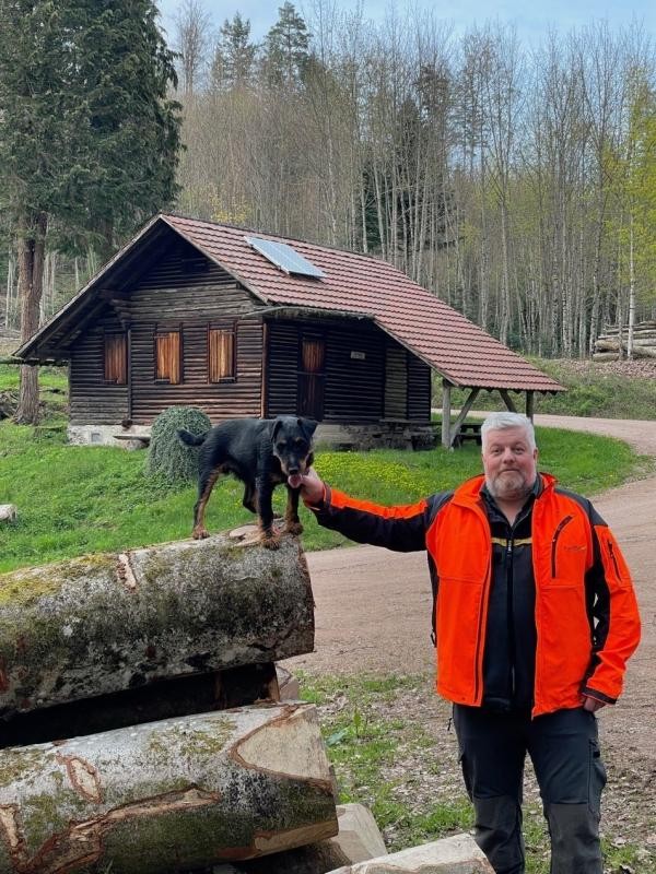 Vor einer Waldhütte steht an einem Waldweg neben einem Baumstammstapel ein Mann mit weißgrauen Haaren und Bart. Er hält die Hand an einen kleinen schwarzbraunen Hund, der auf dem Baumstammstapel steht.