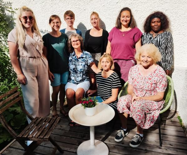 9 Frauen stehen und sitzen der Kamera zugewandt nebeneinander und lächeln.