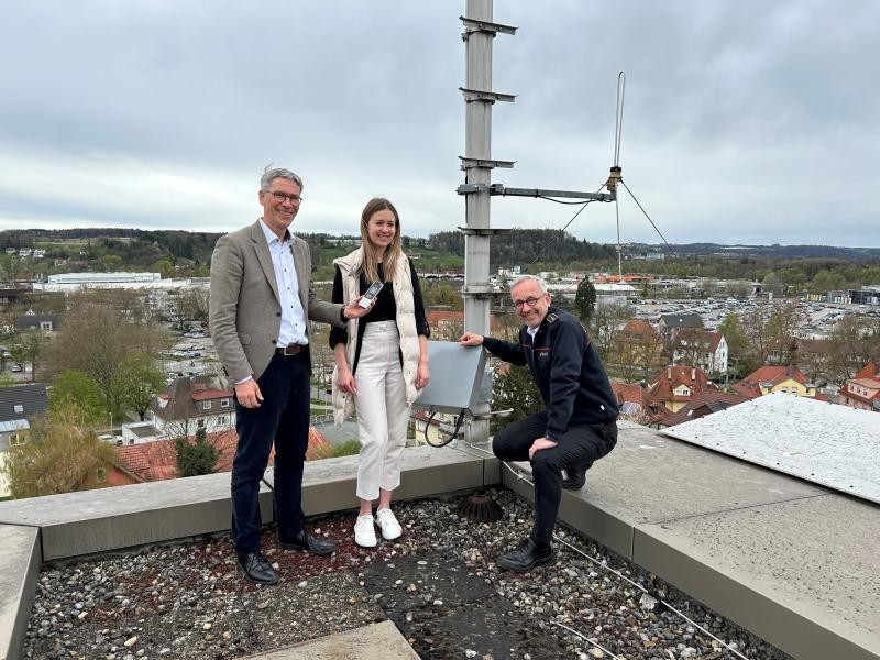 Eine Frau sowie zwei Männer stehen auf einer Dachterasse neben einen neu istallierten Satellit