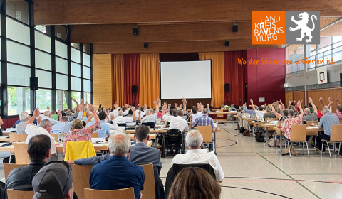 in einer Gemeinde-Mehrzweckhalle sitzen Menschen an Tischreihen und heben jeweils eine Hand zur Abstimmung nach oben.