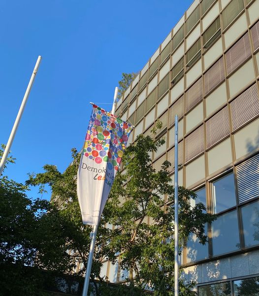 vor einem hohen Gebäude ein Fahnenmast mit einer rechteckigen Flagge, die im oberen Teil mit bunten Farbpunkten versehen ist. Die untere Hälfte ist weiß gehalten. Die Flagge trägt den Schriftzug "Demokratie leben!".