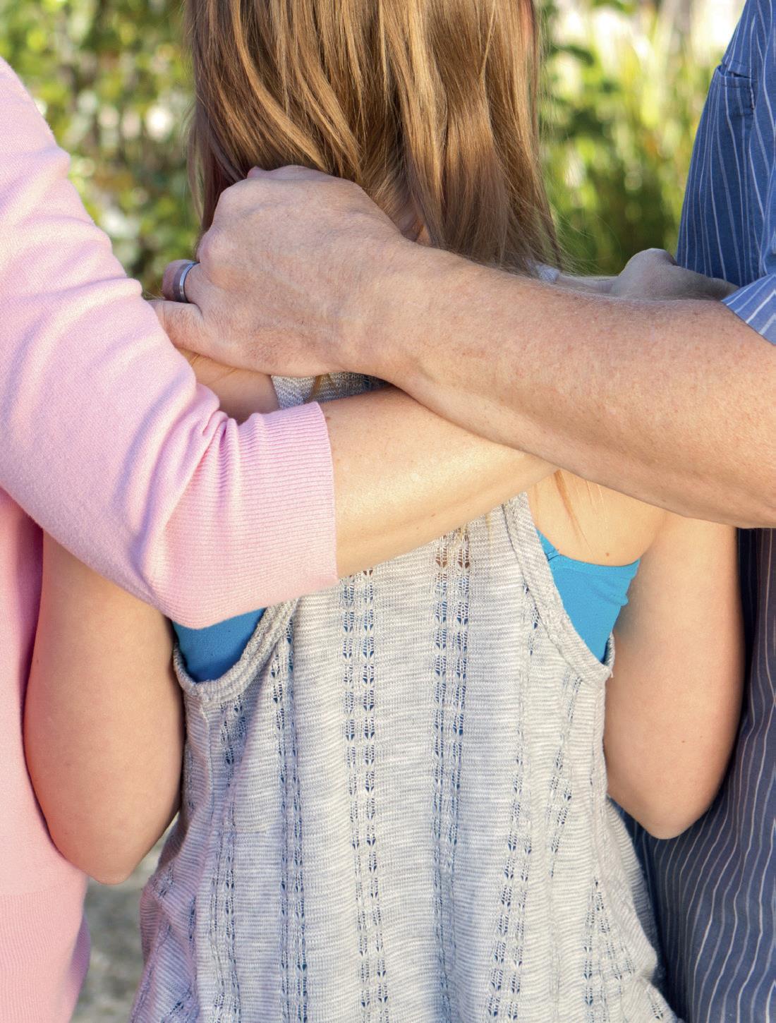 Das Bild zeigt im Zentrum die Rückseite eines weiblichen Kindes mit blonden Haaren. An seiner linken und rechten Seite wird es von jeweils einem Arm einer weiblichen erwachsenen und einer männlichen erwachsenen Person umfasst.