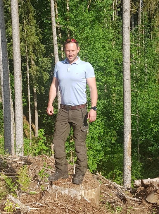 Ein Mann mit Poloshirt sowie Arbeitshose und -schuhe steht im Wald und blickt in die Kamera